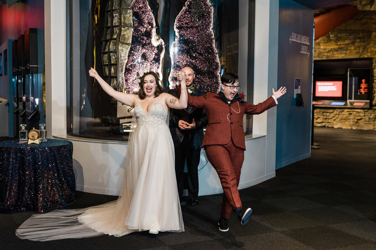 Two brides face their guests and cheer enthusiastically right after their wedding ceremony at the Perot Museum in Dallas, Texas. Both brides are cheering with arms raised high up in the air in celebration. The bride on the left is wearing a long, white dress while the bride on the right is wearing a burgundy suit. Behind them is their officiant and behind him is a giant purple geode in a display case.