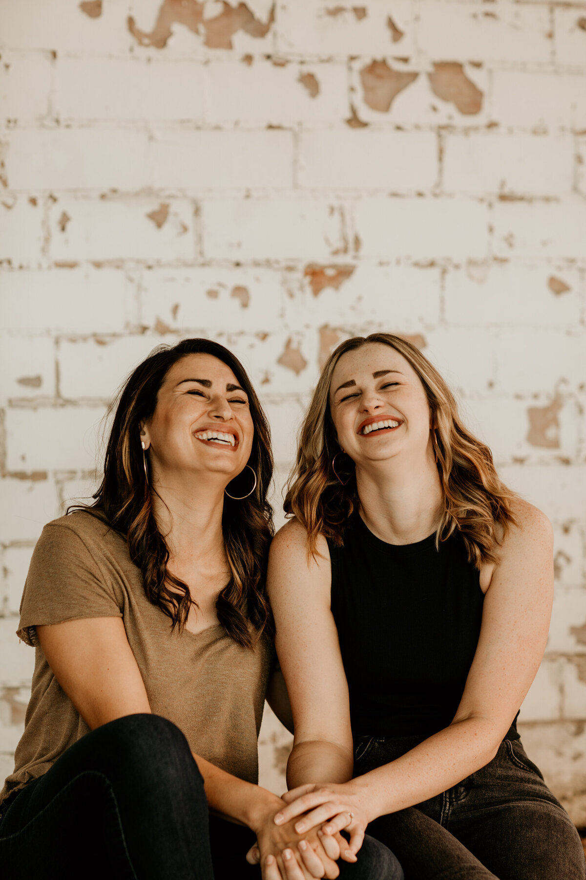 same sex couple laughing  and sitting together in front of a white brick wall