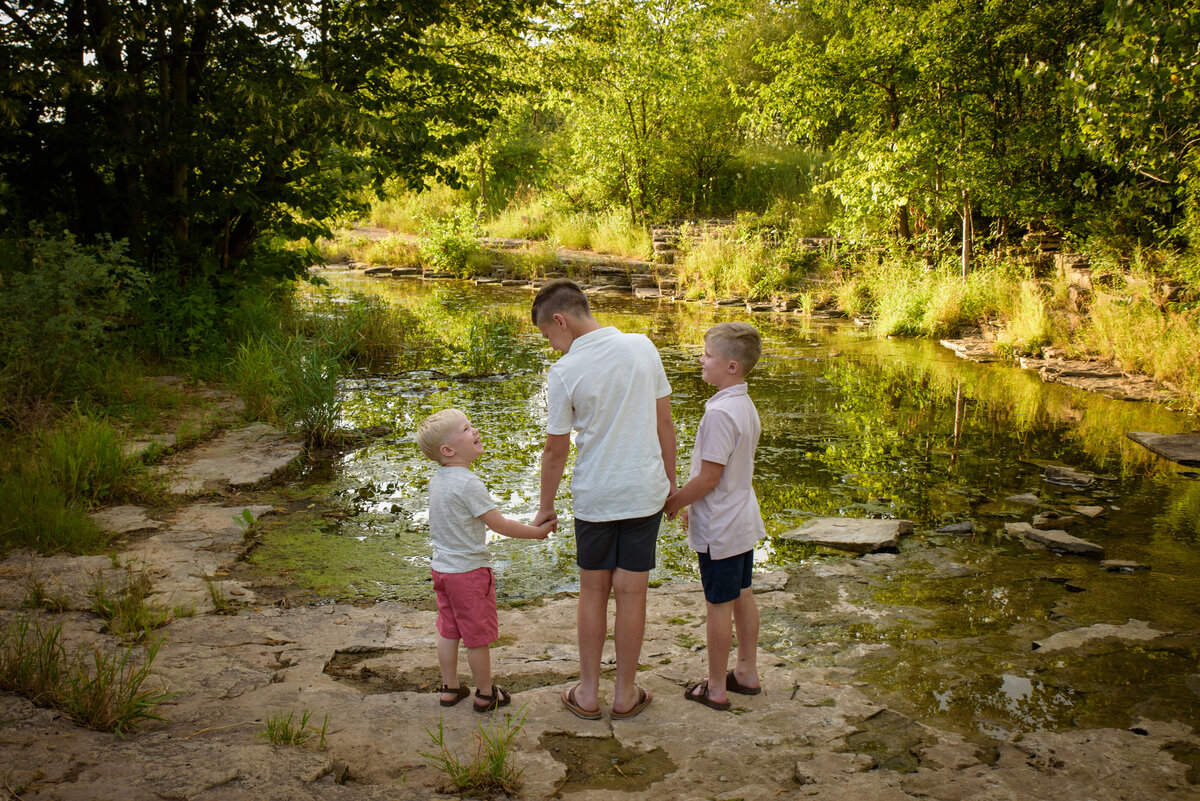 Sibling portrait of three young boys holding hands looking away from the camera near creek at Fonferek Glen County Park near Green Bay, Wisconsin