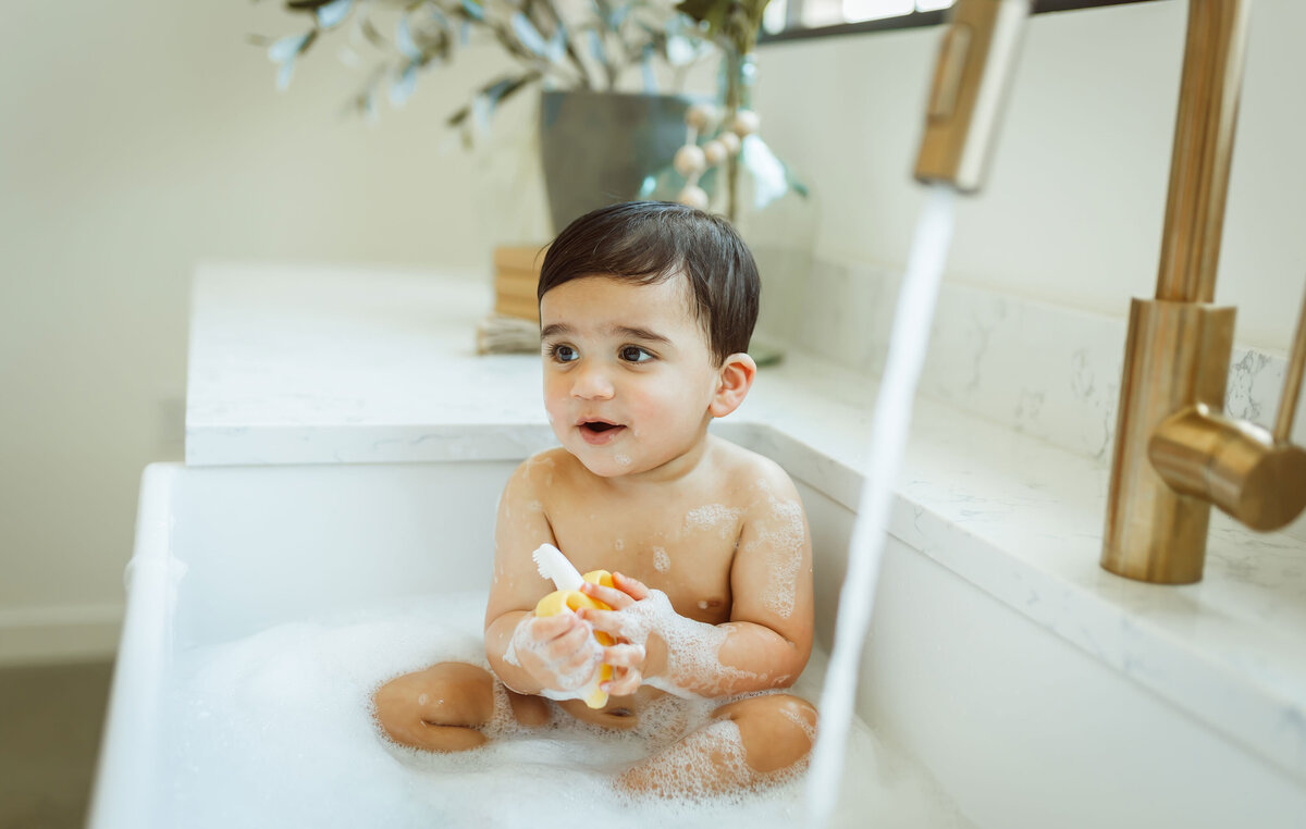 Young boy taking a bath in the kitchen sink with bubbles.