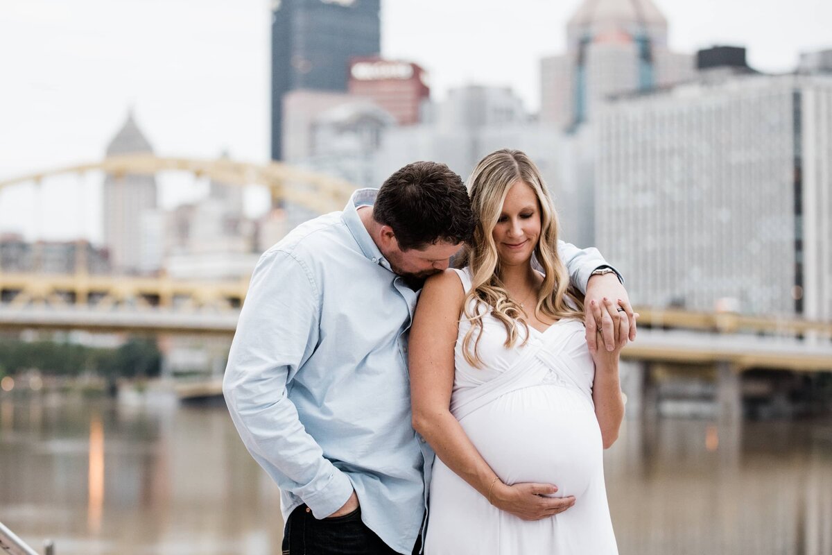 Couple sharing an affectionate moment by the riverfront with a city skyline in the background during their maternity photography session.
