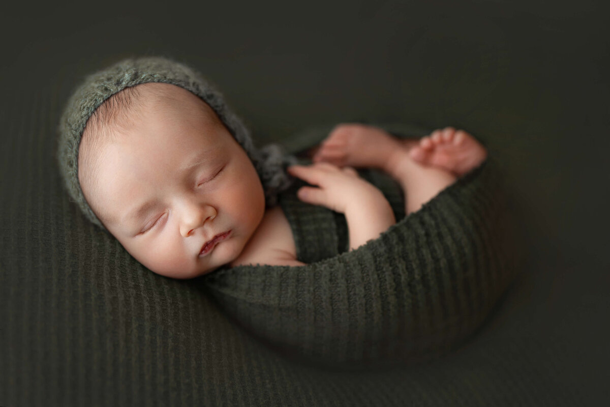 newborn baby boy in a green bonnet and wrap laying on his back  in a swaddle on a green backdrop at his newbonr photography session with a photographer at a loudoun county va photo studio