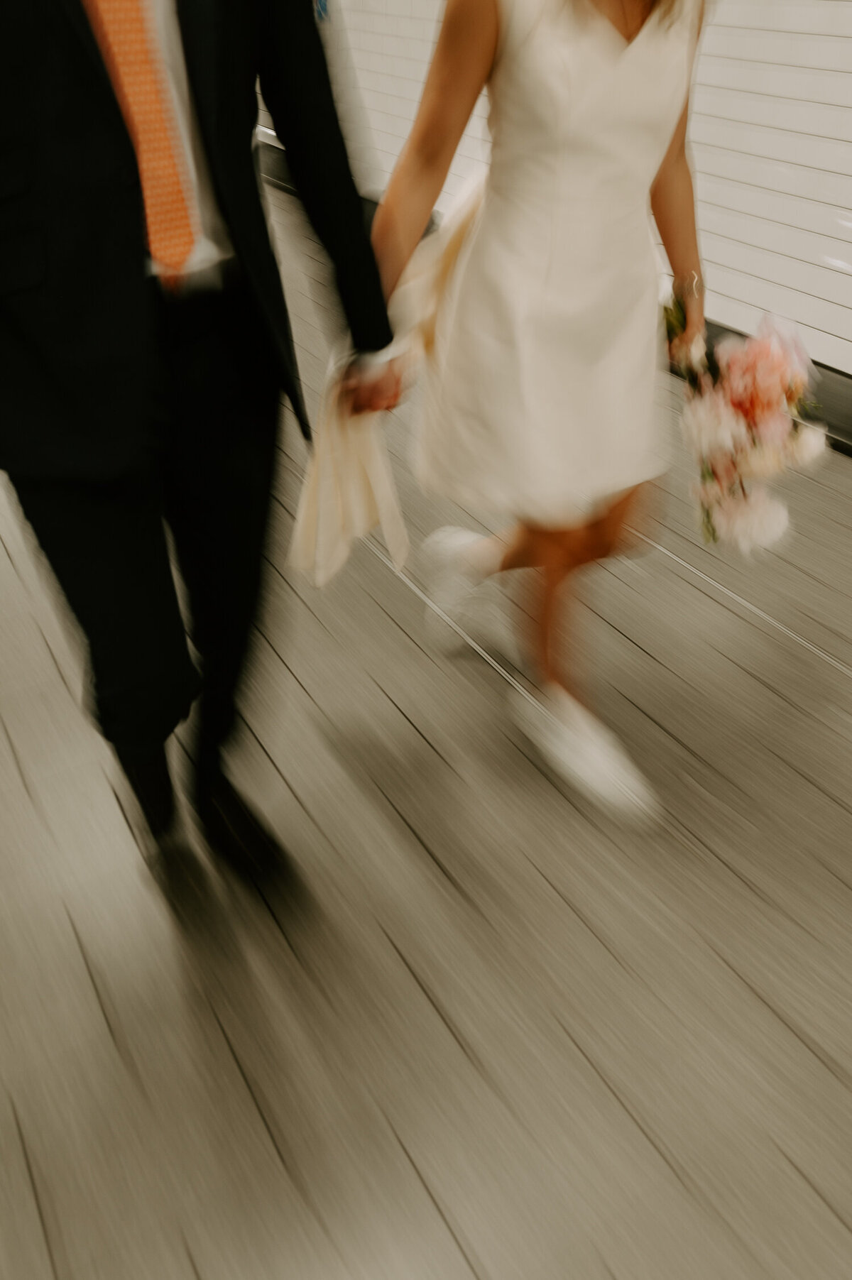 A slow shutter photo of a bride and groom walking through a London tube station, the bride is wearing Prada trainers and has a short wedding dress. Her flowers are pink and cream.