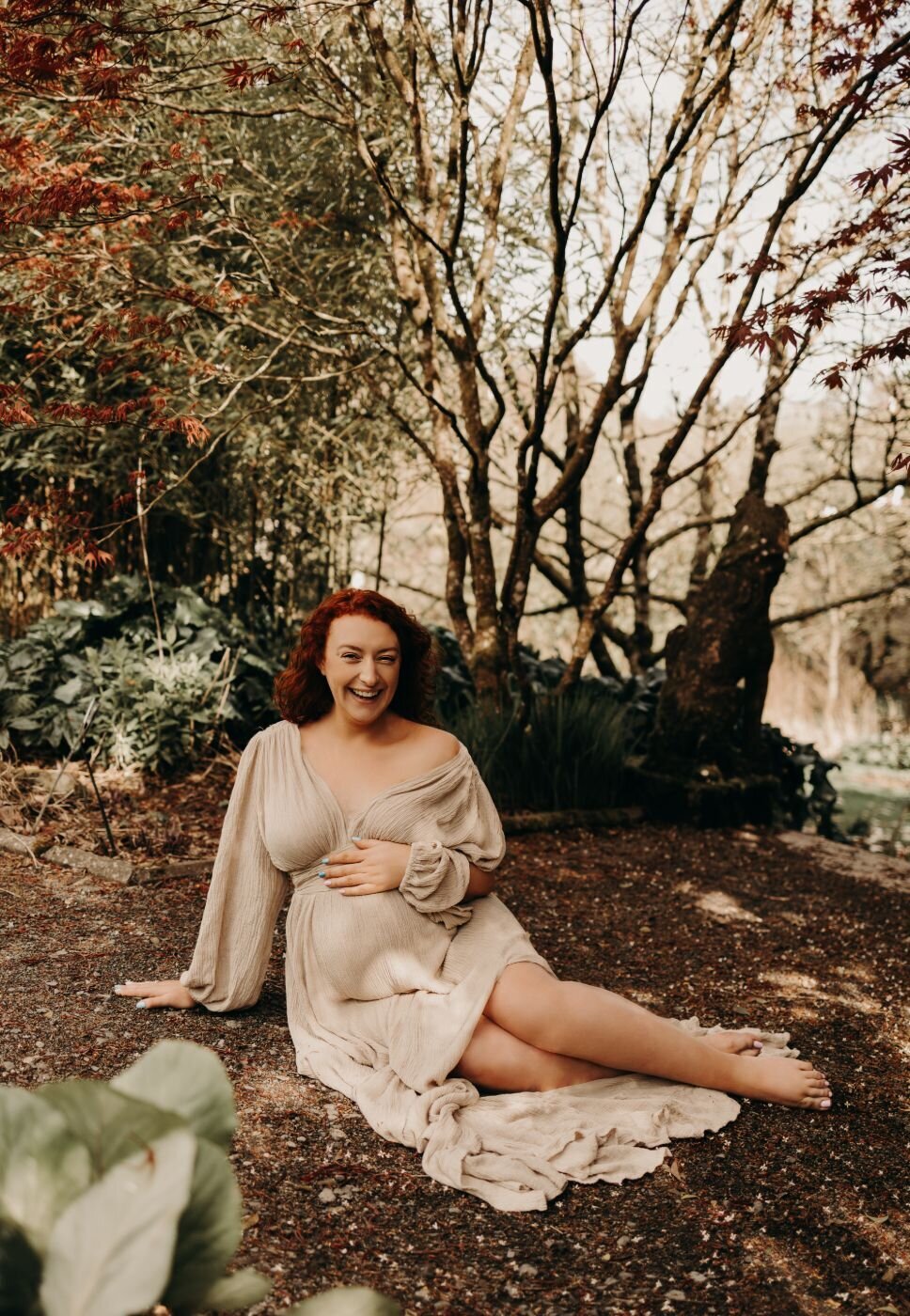 Laughing pregnant woman with red hair in a flowing gown during an outdoor maternity shoot