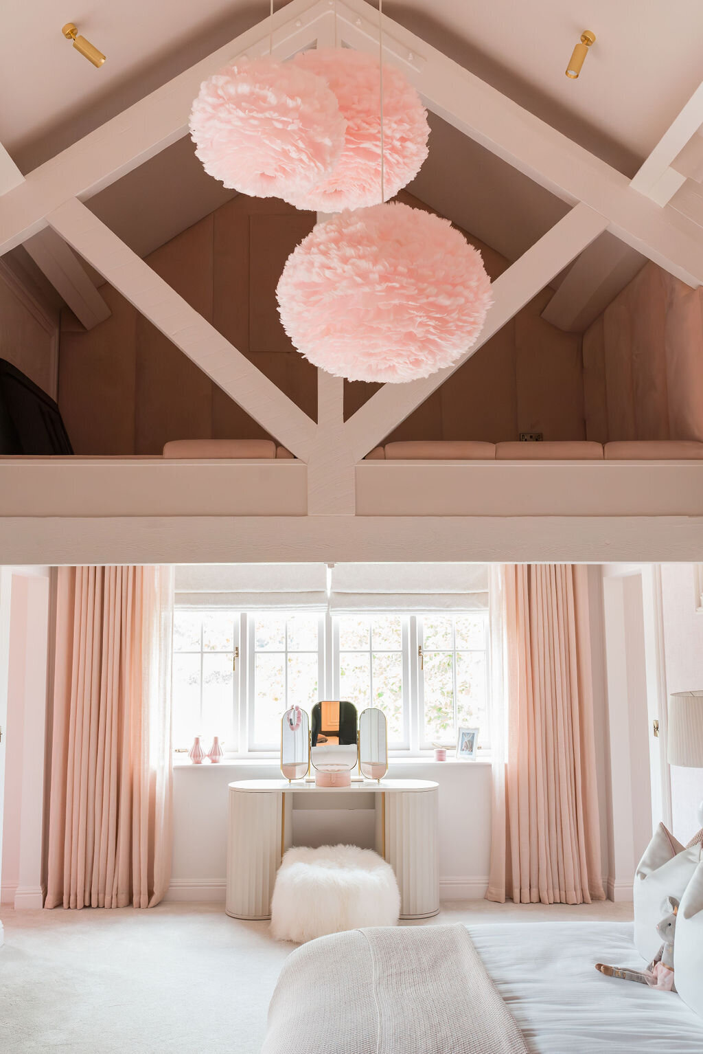 A cozy bedroom with pastel pink decor features a loft with wooden beams overhead. The room has two large, fluffy pink chandeliers, floor-length pink curtains, and a vanity with a large mirror and a white stool.