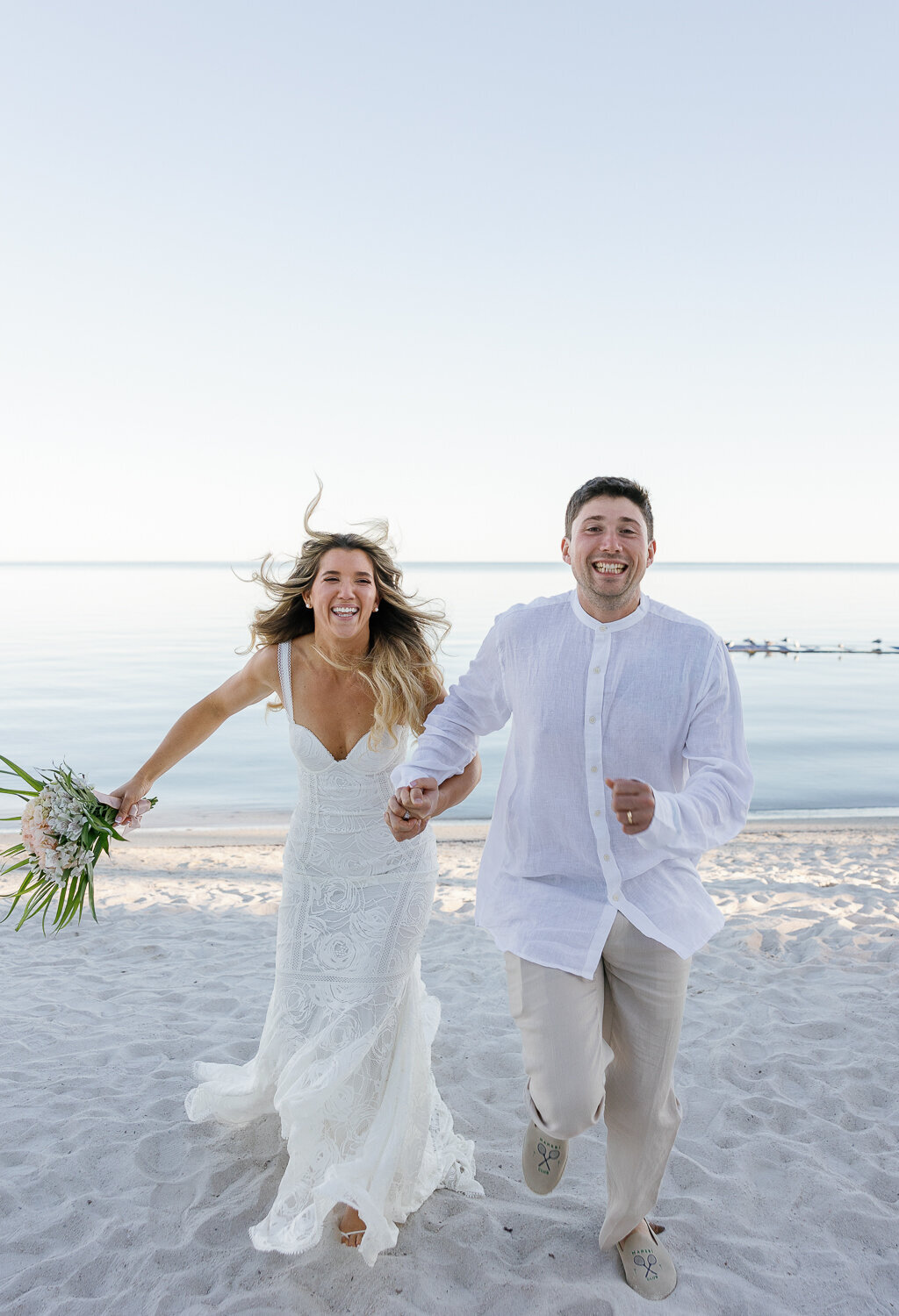 Newlyweds Laura and Abraham joyfully running along the white sand beach at their Miami destination wedding.