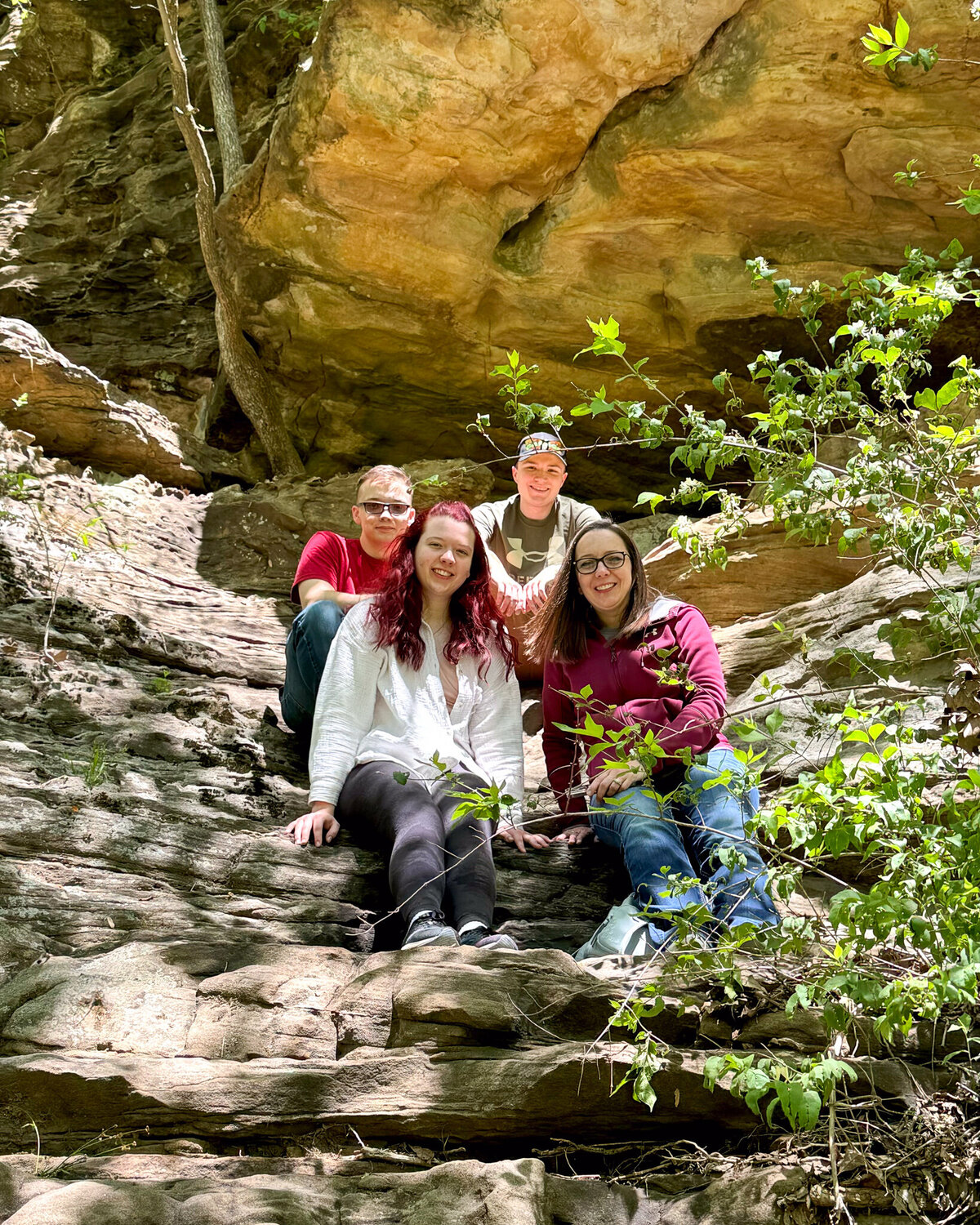 a mom with her 3 adult kids sitting next to each other on a rock formation with green vegetation around them smiling at the camera