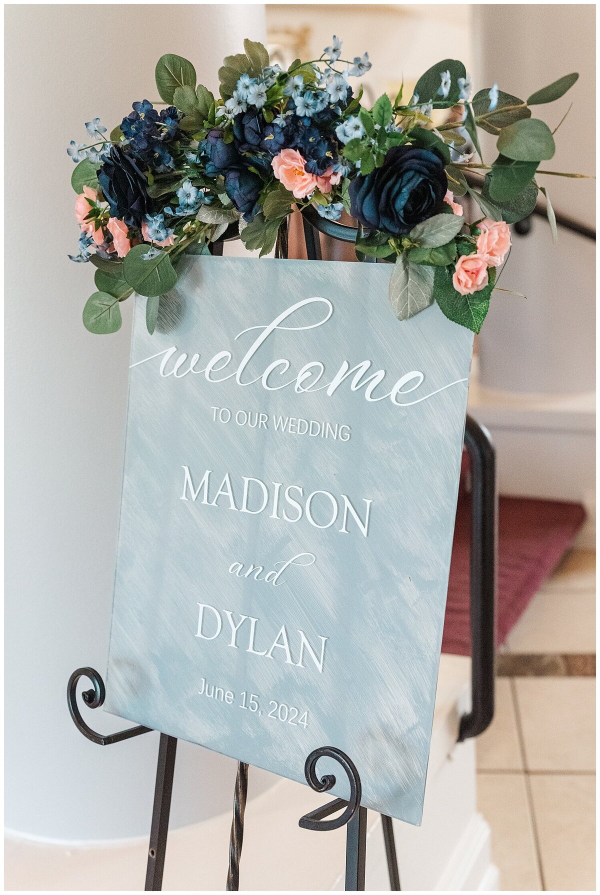detail shot of a dusty blue welcome sign with navy, light blue, and pink flowers surrounded by greenery on top  of the sign for Madison and Dylan at their wedding at Chateaux at Fox meadows captured by denver wedding photographer