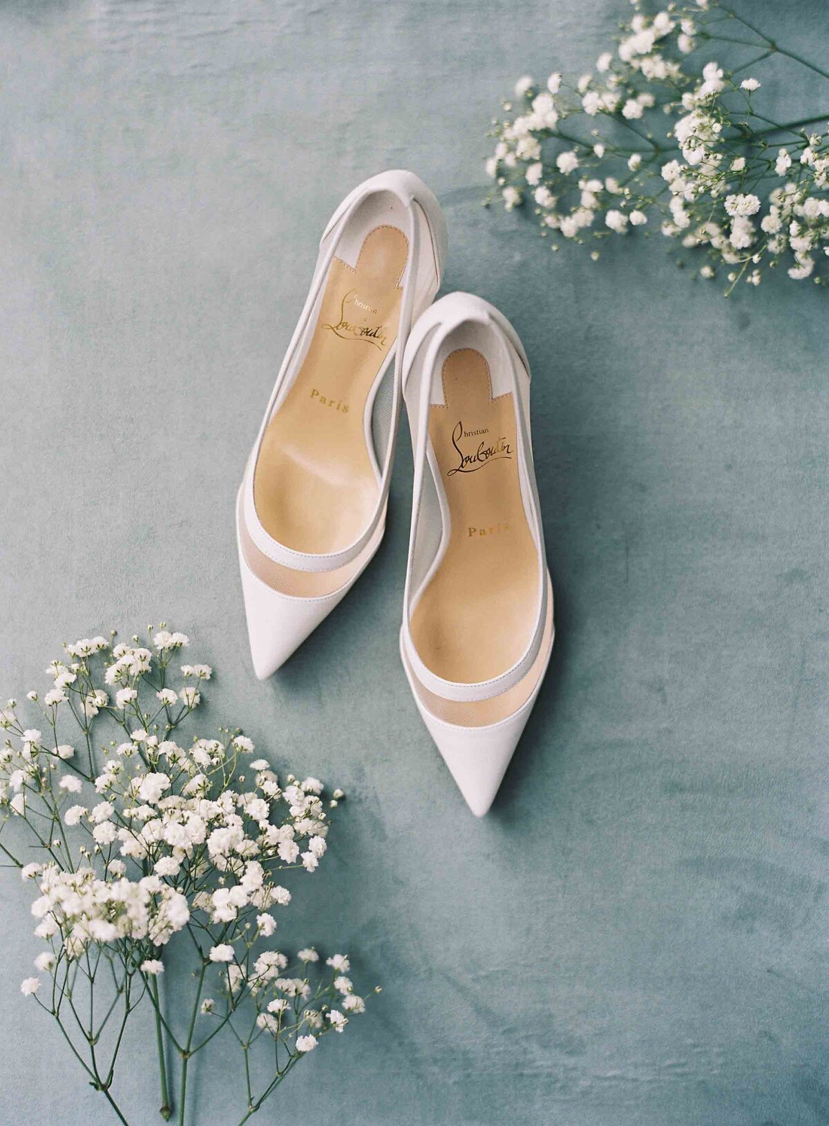 christian louboutin white birdal heels on blue velvet flat lay surface with babies breath flowers surrounding them