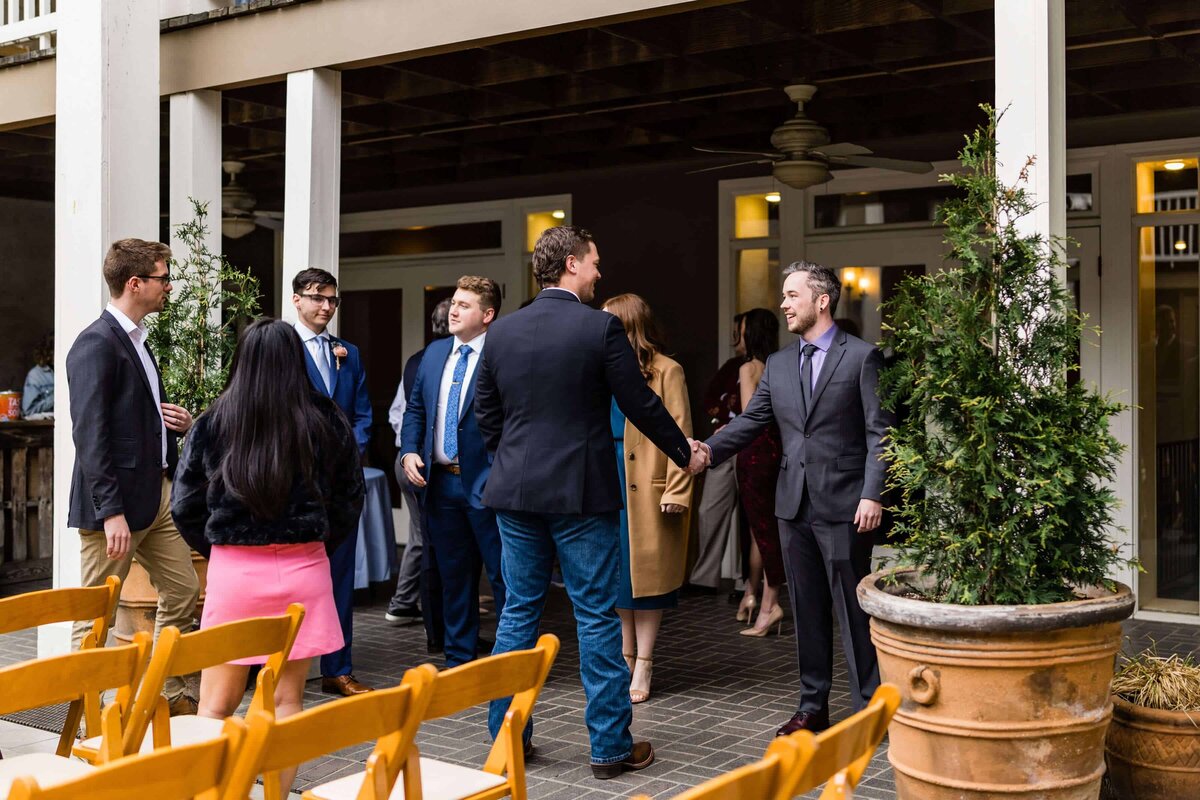 A group of people dressed in formal attire stand in a courtyard near wood chairs; two men in suits shake hands while others converse around them, with potted plants in the foreground.