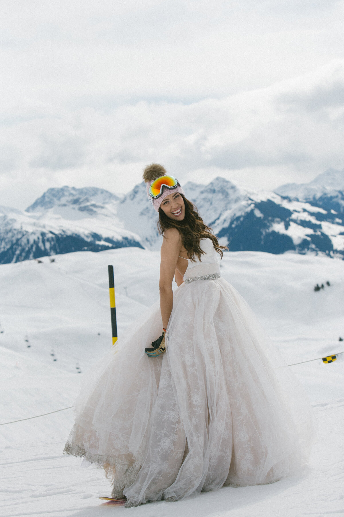 Bride in wedding dress and ski goggles smiling on snowy mountaintop in Kitzbühel – playful winter wedding portrait by Studio Rose