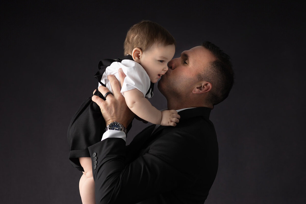DFW milestone photographer captures a touching moment of a father in a black suit kissing his baby boy on the cheek, both dressed formally against a dark studio backdrop.