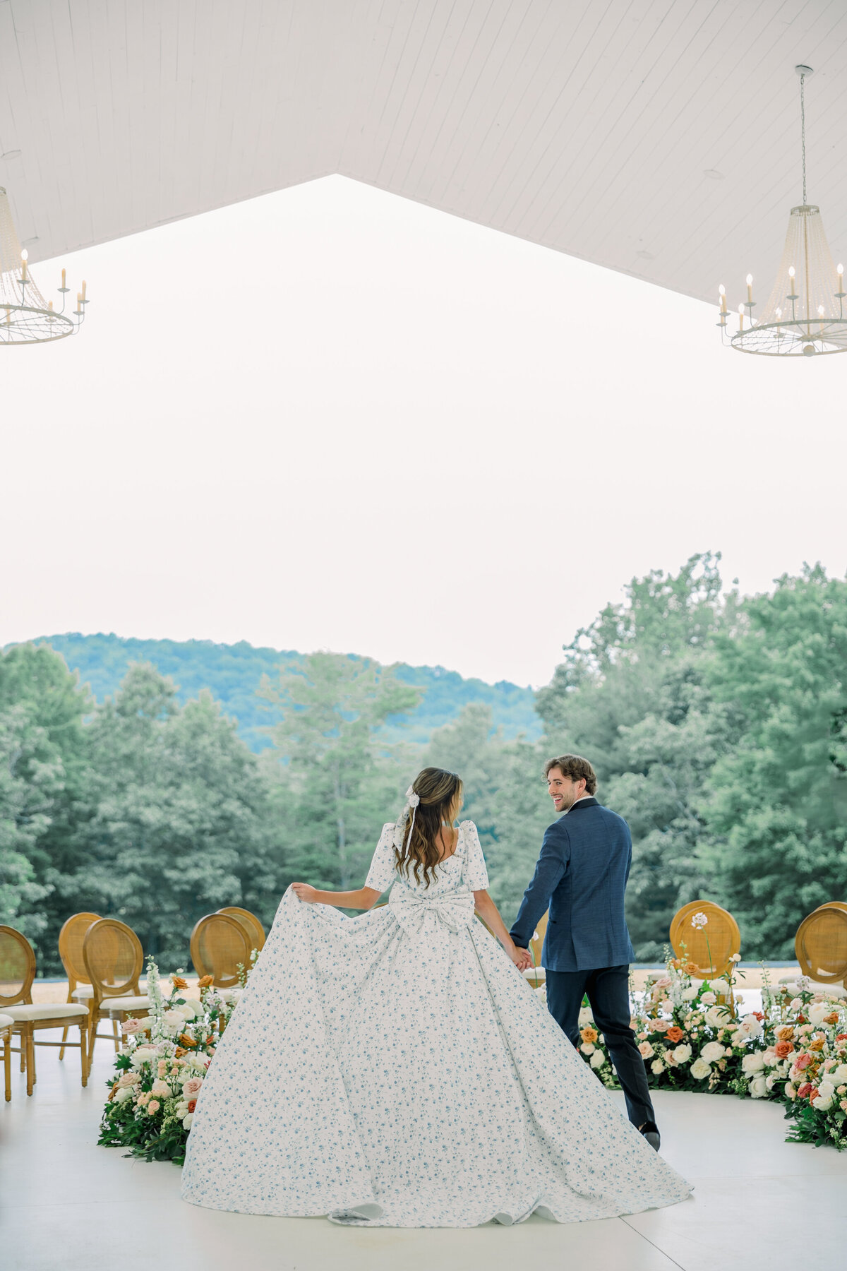 Ivy Rose Barn | A bride in a vintage-inspired floral gown with puff sleeves and a bow walks hand in hand with her groom in a navy tuxedo towards their floral-filled ceremony site in the pavilion of the Ivy Rose Barn.