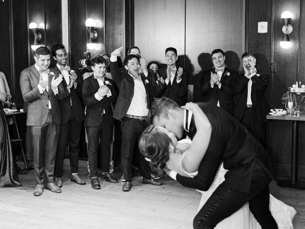 During their first dance the groom dips his bride back as he kisses her and their friends and family cheer loudly