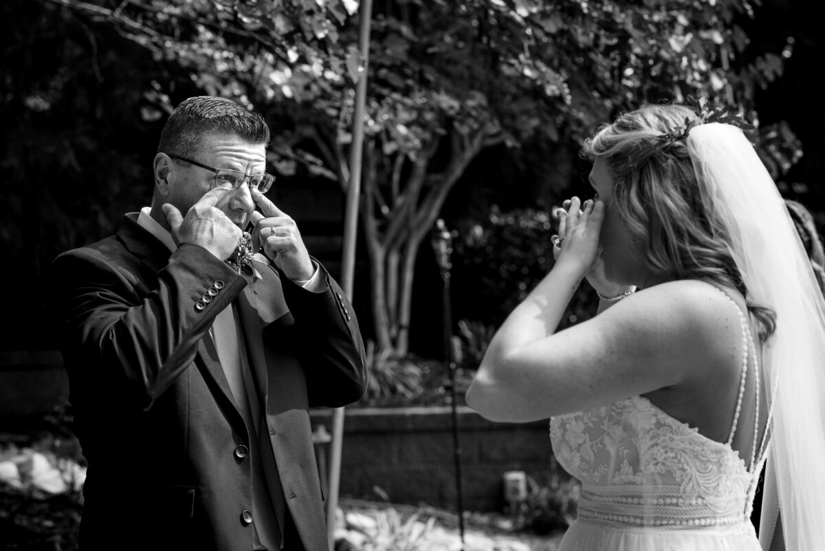 Black and white image of a father-daughter first look outside where both are crying and wiping tears by Charlotte wedding photographers DeLong Photography