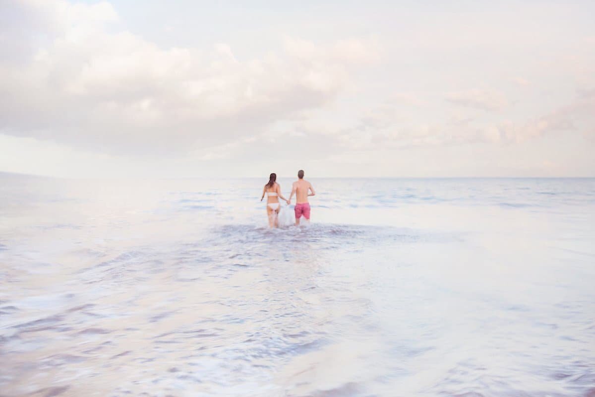 Couple in swimsuits run away from the camera in to the ocean during their beach engagement session on Maui