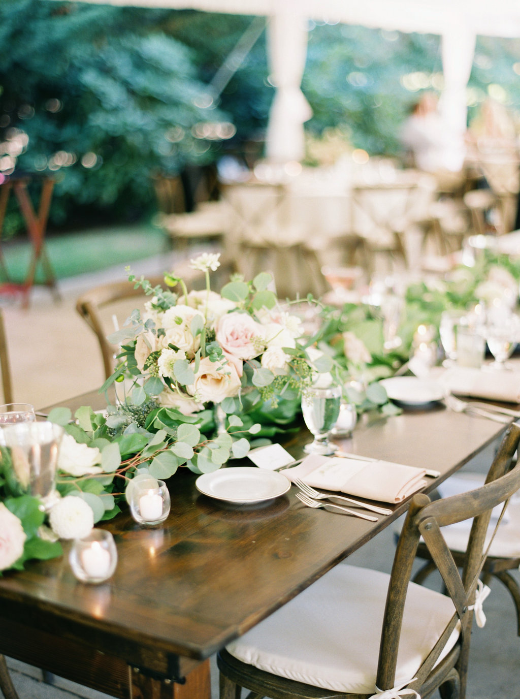 Long wooden wedding reception table, greenery runner with roses tucked into it make for an easy and beautiful centerpiece.