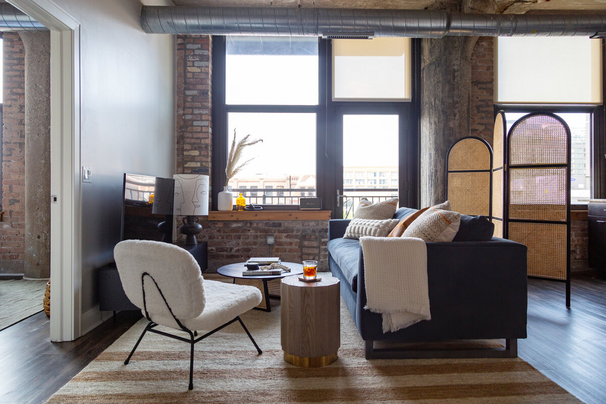 Industrial loft featuring brick walls and concrete columns with blue velvet sofa, arched cane divider screen, striped jute rug, and furry white lounge chair