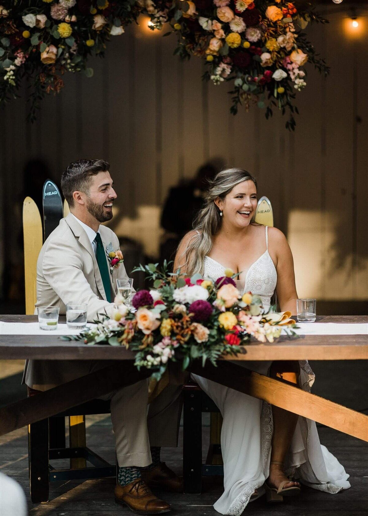 Bride and groom sit at their sweetheart table with a large floral arrangement.