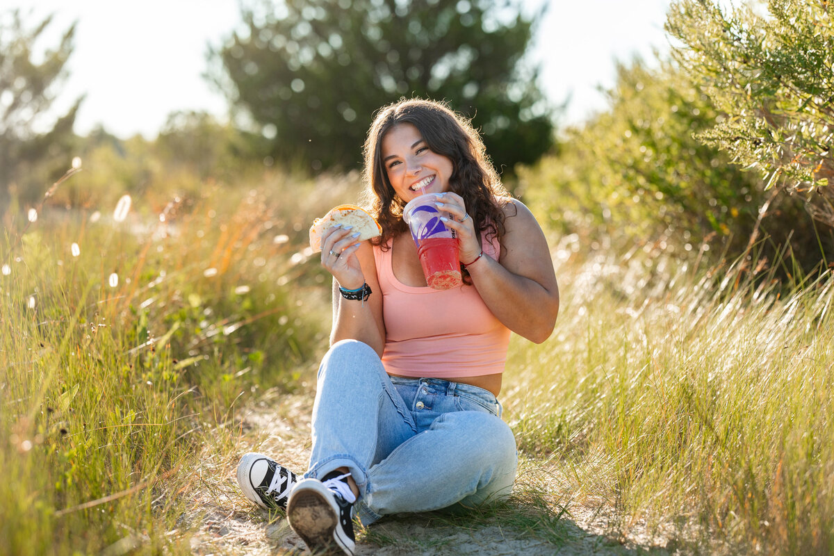A high school senior lounges in the golden light of a sunlight field in Virginia, laughing as she holds a Taco Bell taco and Baja Blast.  Dressed in a peach tank top, jeans and sneakers, she radiates confidence, humor, and authentic joy in this playful Brooke Tucker Photography session.