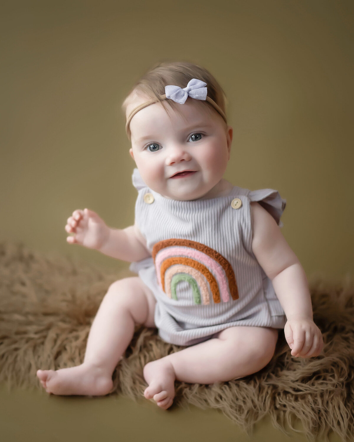 Baby in a striped outfit with a rainbow design sitting on a fuzzy rug.