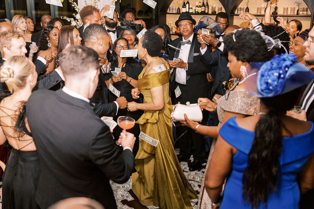The groom dances with an elegantly dressed guest in a gold gown, surrounded by family and friends in formal attire.