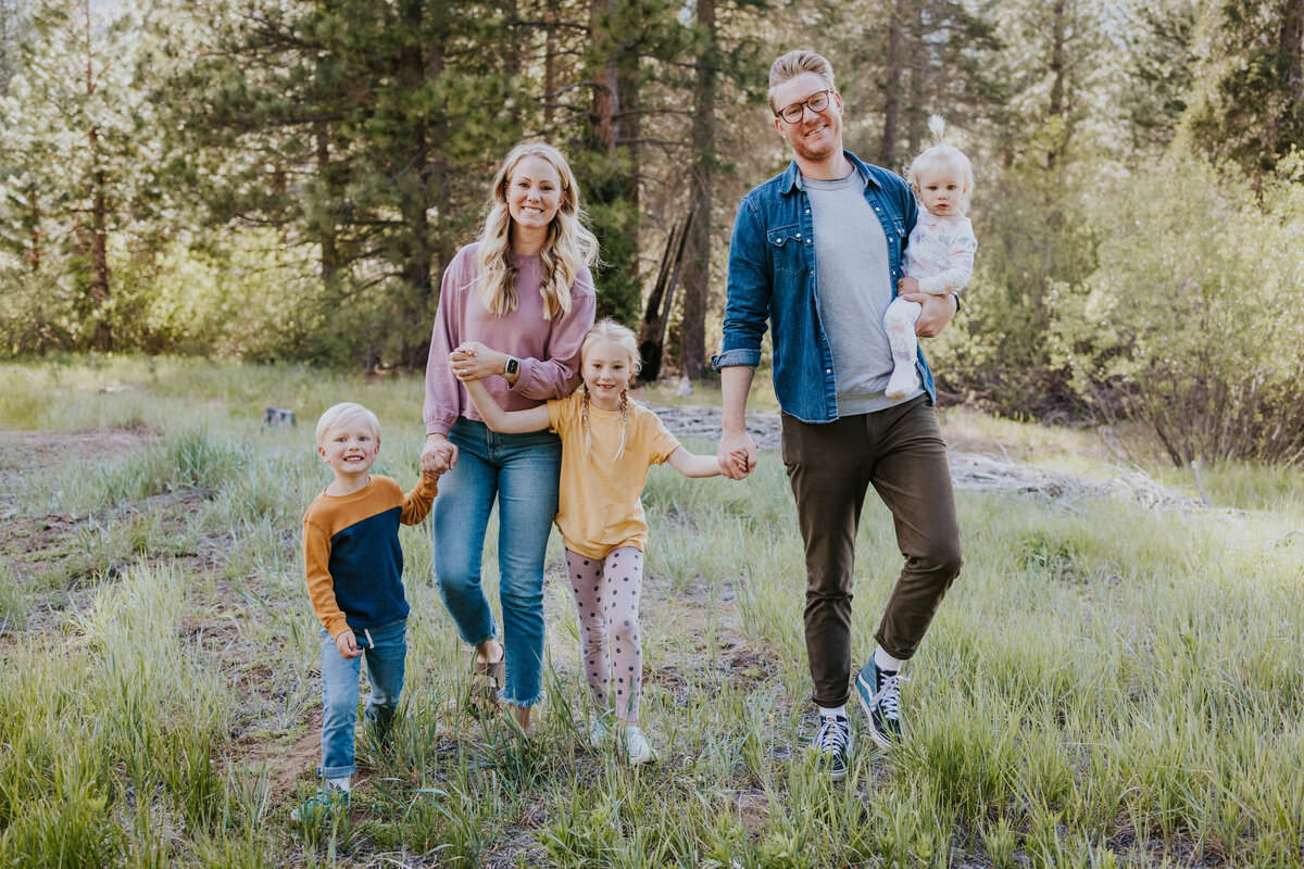Family of five hold hands and walks through forest meadow while smiling at camera.
