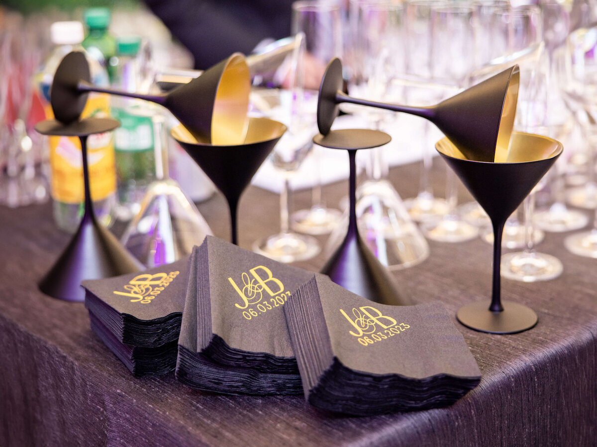 A collection of sleek, black martini glasses with gold interiors are displayed on a counter alongside a stack of personalized black napkins featuring the initials J & B and the date 06.03.2023. In the background, empty glassware is visible.