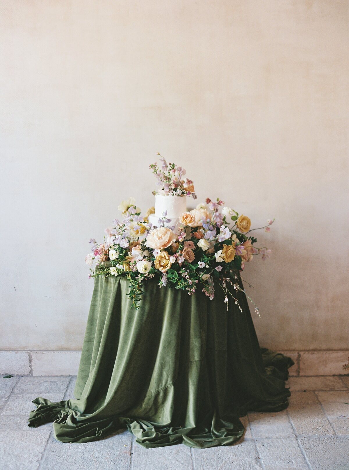 small white wedding cake surrounded by flowers and a dark green table cloth