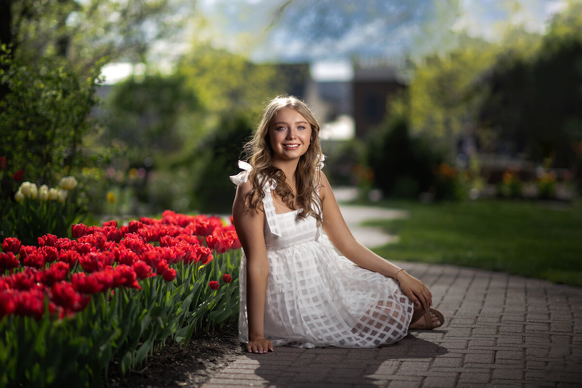 A high school senior in a white dress sits in the sidewalk of a colorful garden smiling for an Iowa Senior Photographer