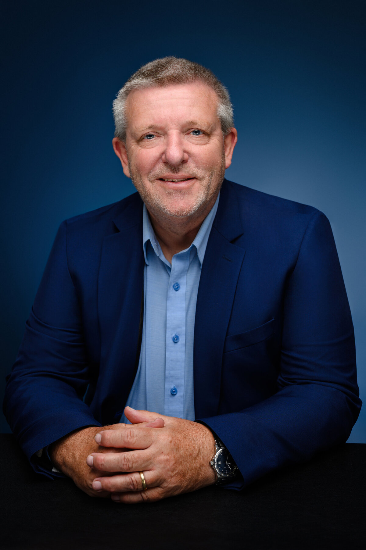 Smiling man in a blue suit and shirt, sitting with his hands clasped