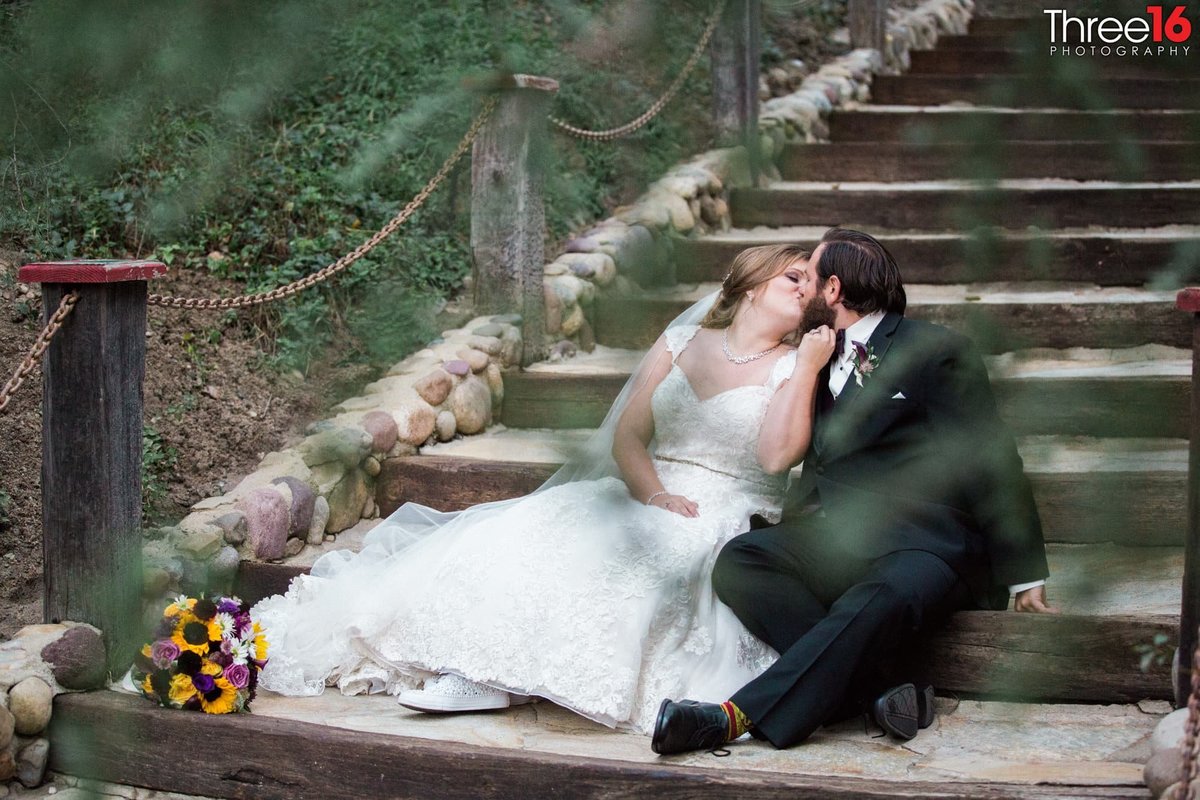 Bride and Groom sit on the outdoor steps share a tender kiss
