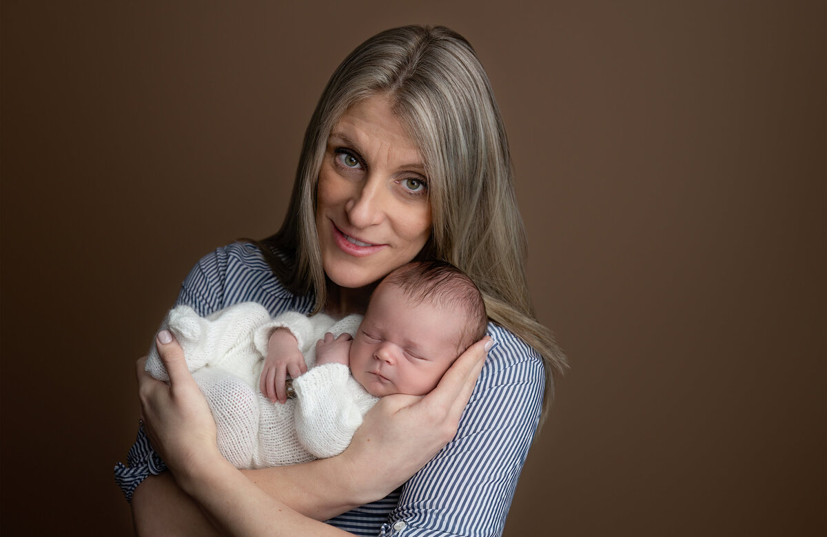 A studio portrait of a mother with soft blonde hair holding her newborn baby in a cozy embrace. Photographed at Meraki Photographic Creations in Paris, Ontario.