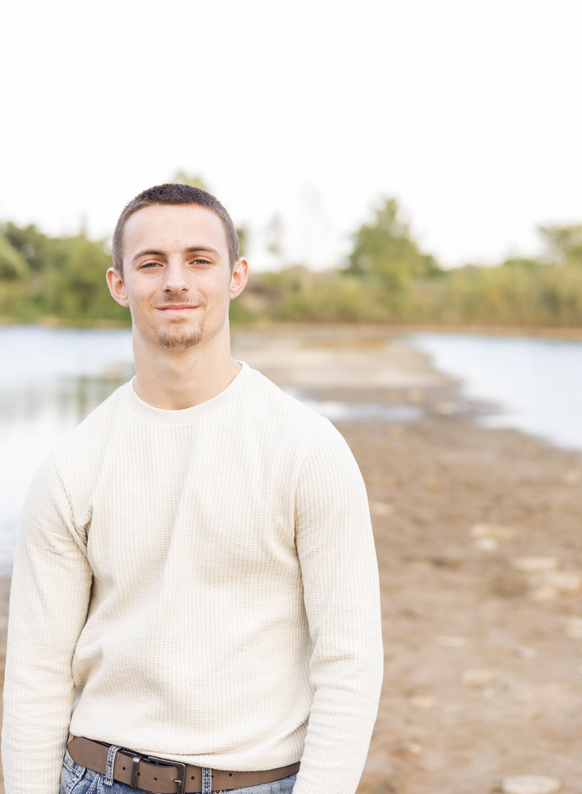 A high school senior male, standing next to a pond, soft smile at the camera, with short brown hair, wearing a light tan long sleeve shirt.