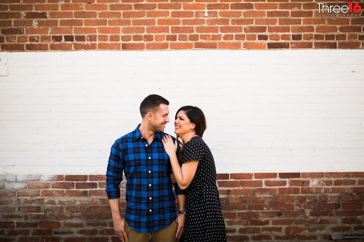 Engaged couple smile at each other in front of a brick wall