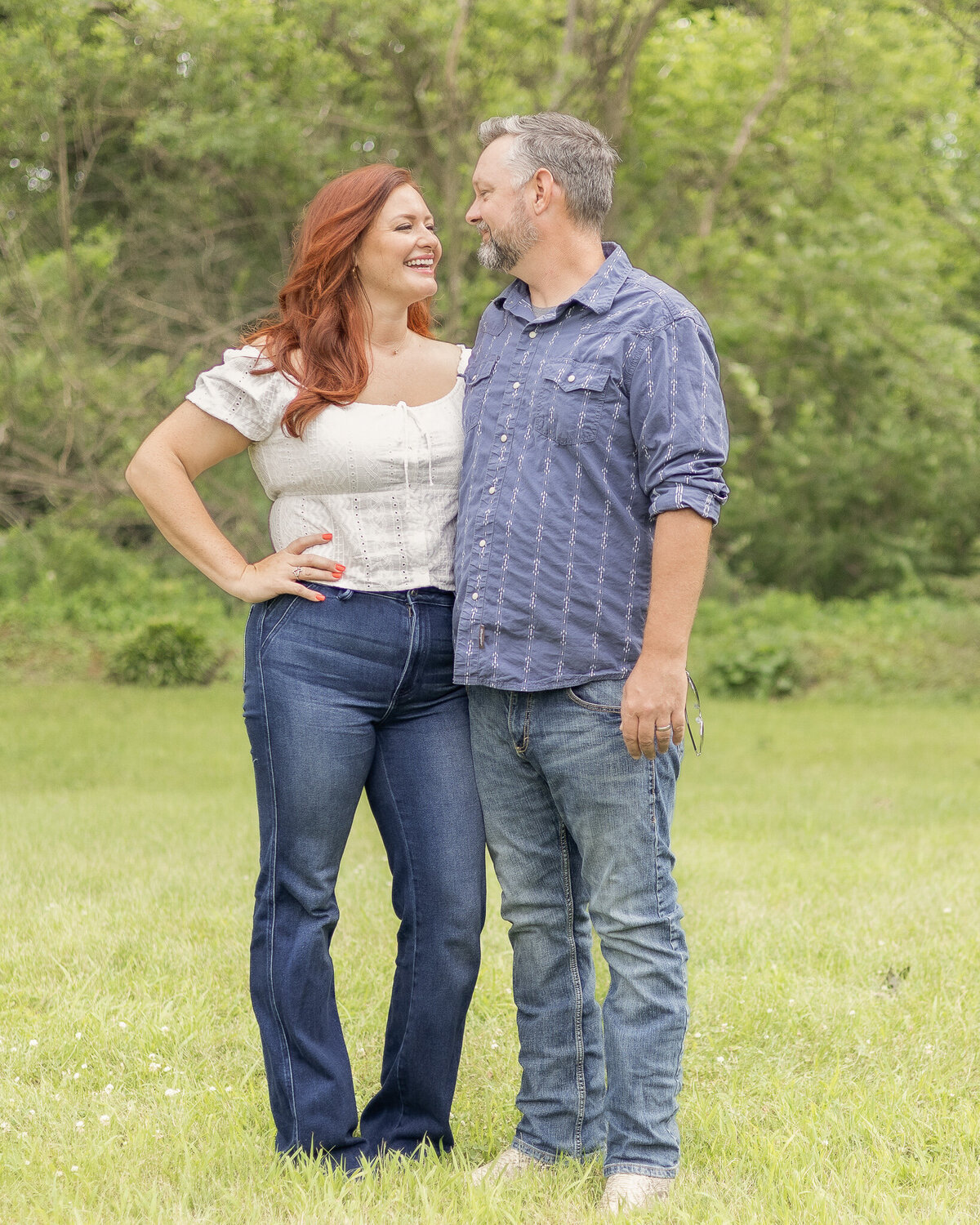 Husband and wife standing in the grass with a tree line behind them.  They are looking at each other laughing.  Wife has a short sleeve shirt on and blue jeans with long red hair.  Husband is looking at her with a pepper hair and beard, wearing a button up shirt with the sleeves rolled up and jeans