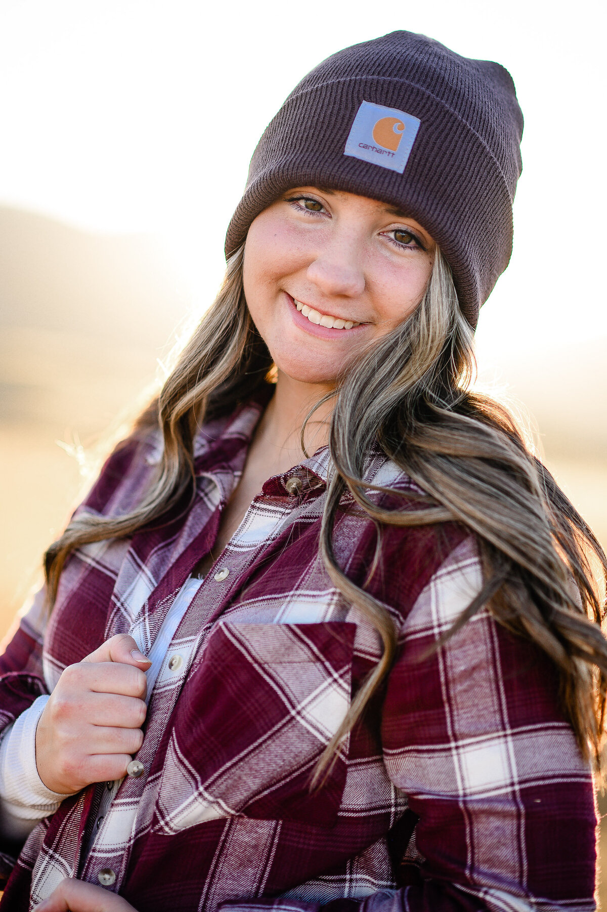 A girl standing in glowy  sunshine in a flannel shirt and carhartt hat smiling for her senior photos