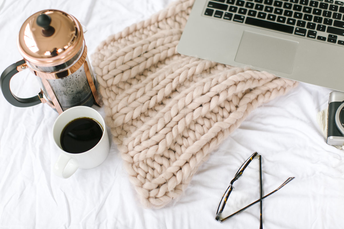 Flatlay of coffee, laptop, pink knitted blanket and glasses on white bedsheet