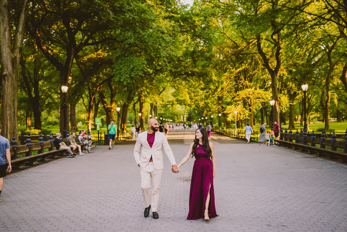 An engagement couple in red formal outfits walking through central park mall