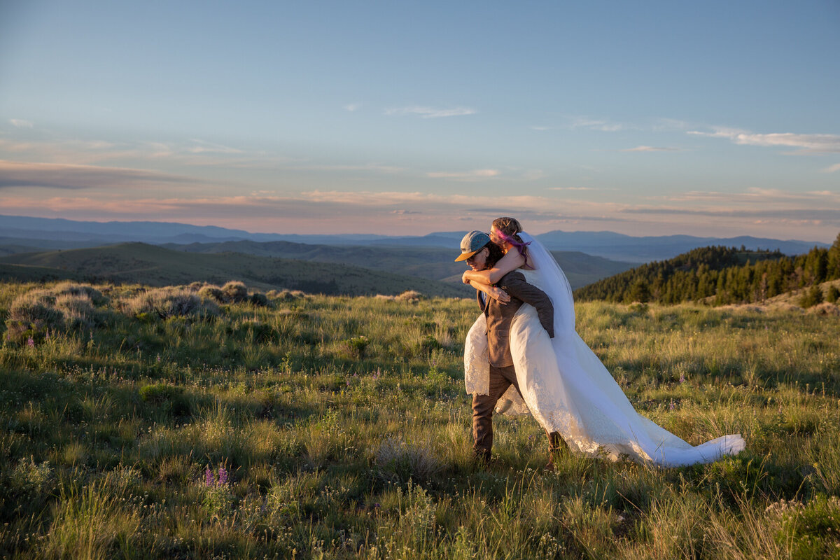 A groom gives his bride a piggy back ride as he walks through a grassy field.