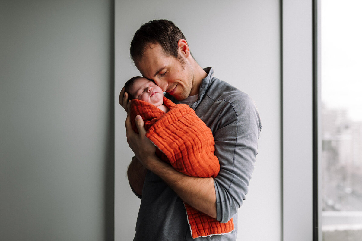 father holding newborn wrapped in orange swaddle up to his face and smiling with his eyes closed