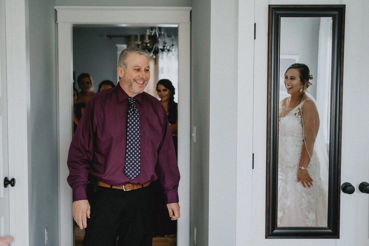 A smiling man in a purple shirt opens a door to a room where a bride, seen in a mirror, is laughing joyfully.