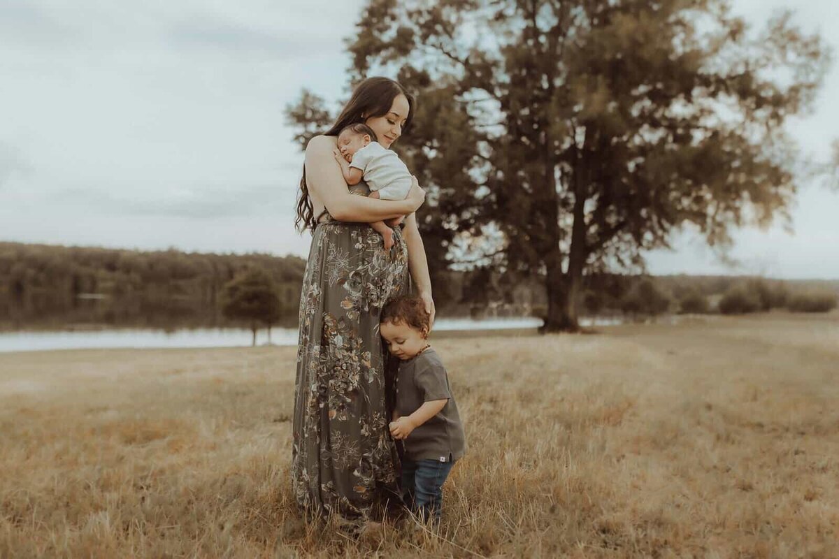 A woman in a long floral dress holds a baby and stands beside a young child in a grassy field, with trees and a lake behind them—an outdoor newborn photography session by Fire Family Photography.
