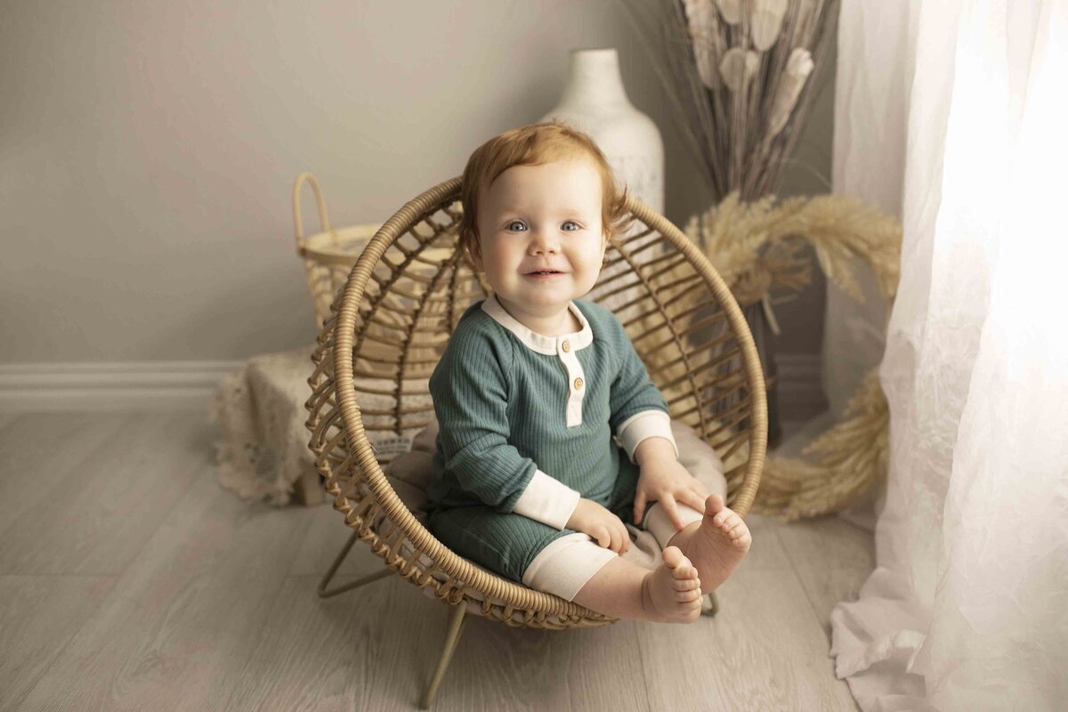 One-year-old birthday boy, dressed for his special day, sitting in a wicker chair with a cheerful expression in the studio