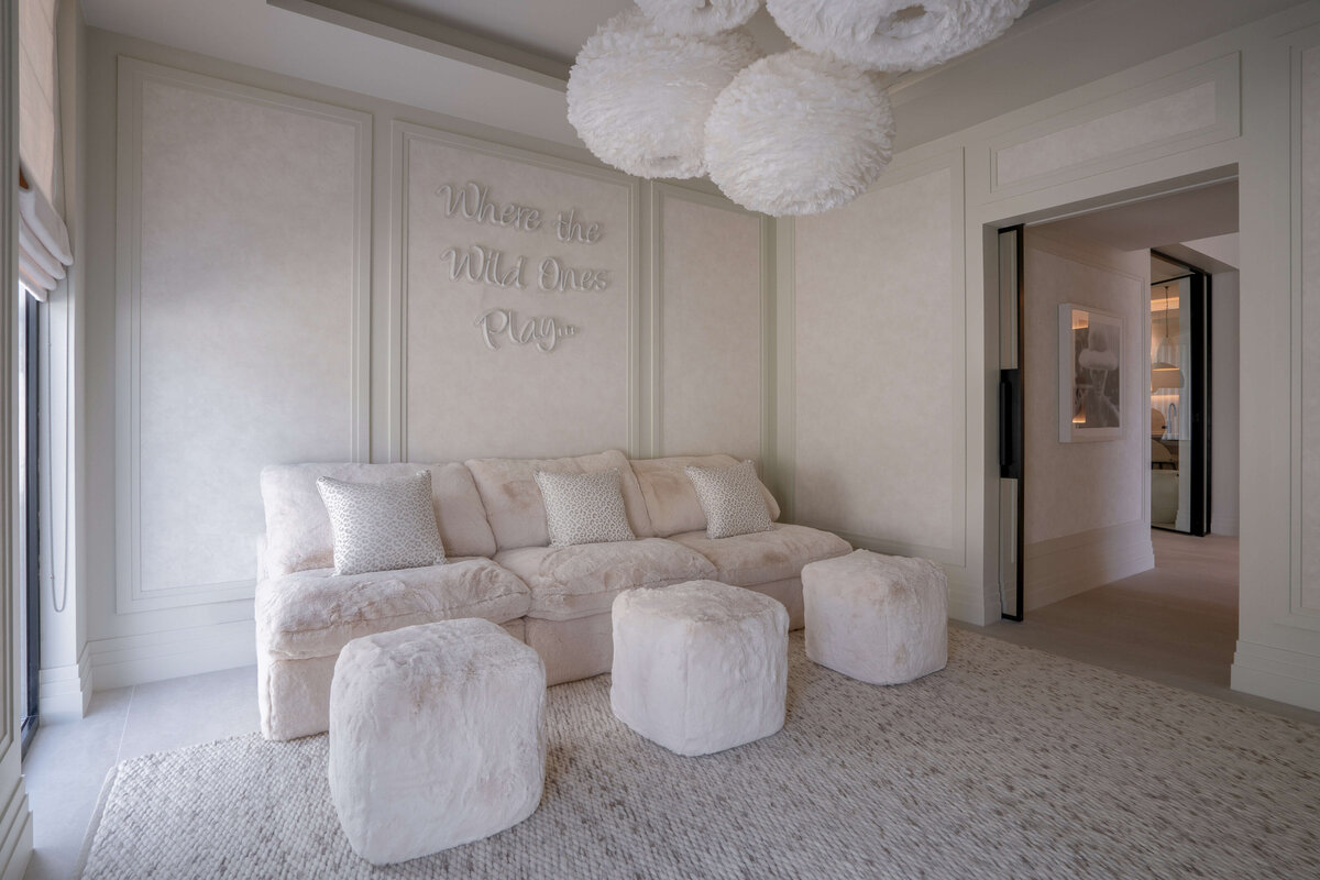 Spacious playroom with a light, airy aesthetic. There is a large, plush tan couch with three fuzzy pouf ottomans. Behind the couch is a neon  sign reading 'Where the Wild Ones Play...'. Above are four white, feathery spherical pendant chandeliers.