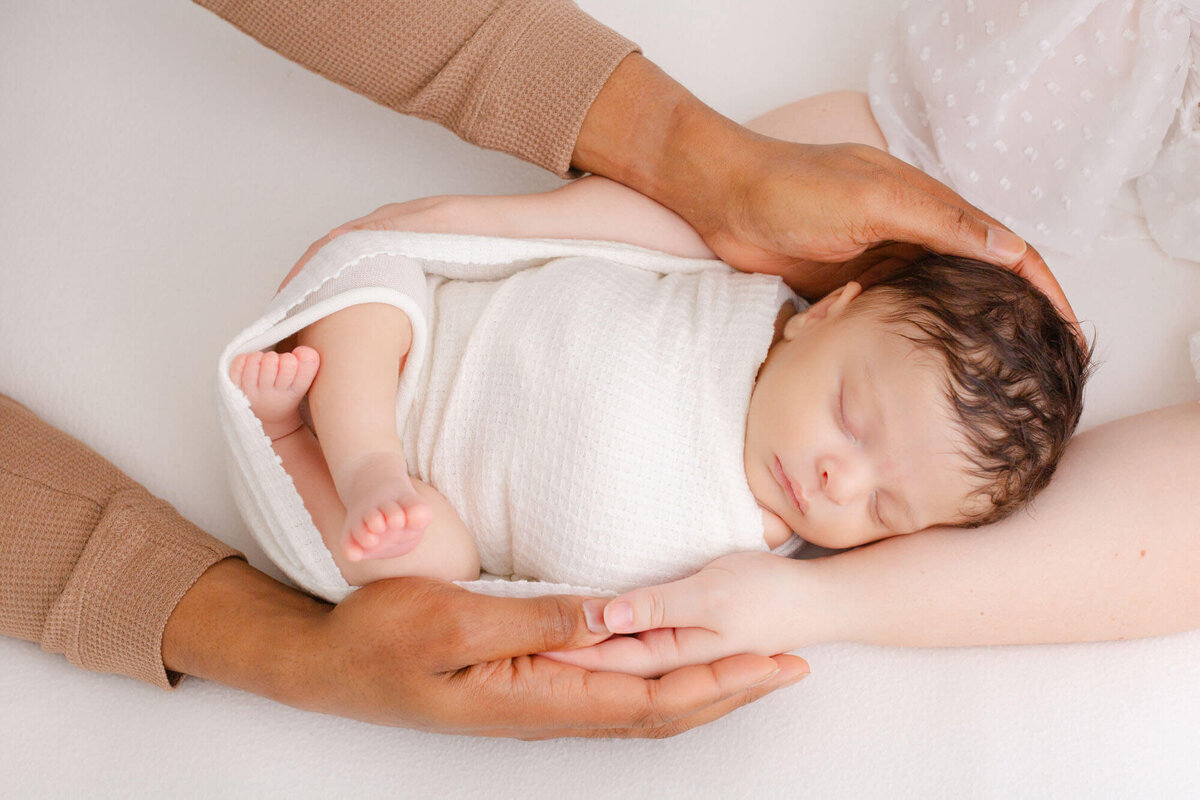 Baby swaddled in white and laying on a white blanket. Mom and Dad are holding baby with both of their arms. You can only view their hands in the image. Portland newborn photoshoot by Ashlie Behm Photography