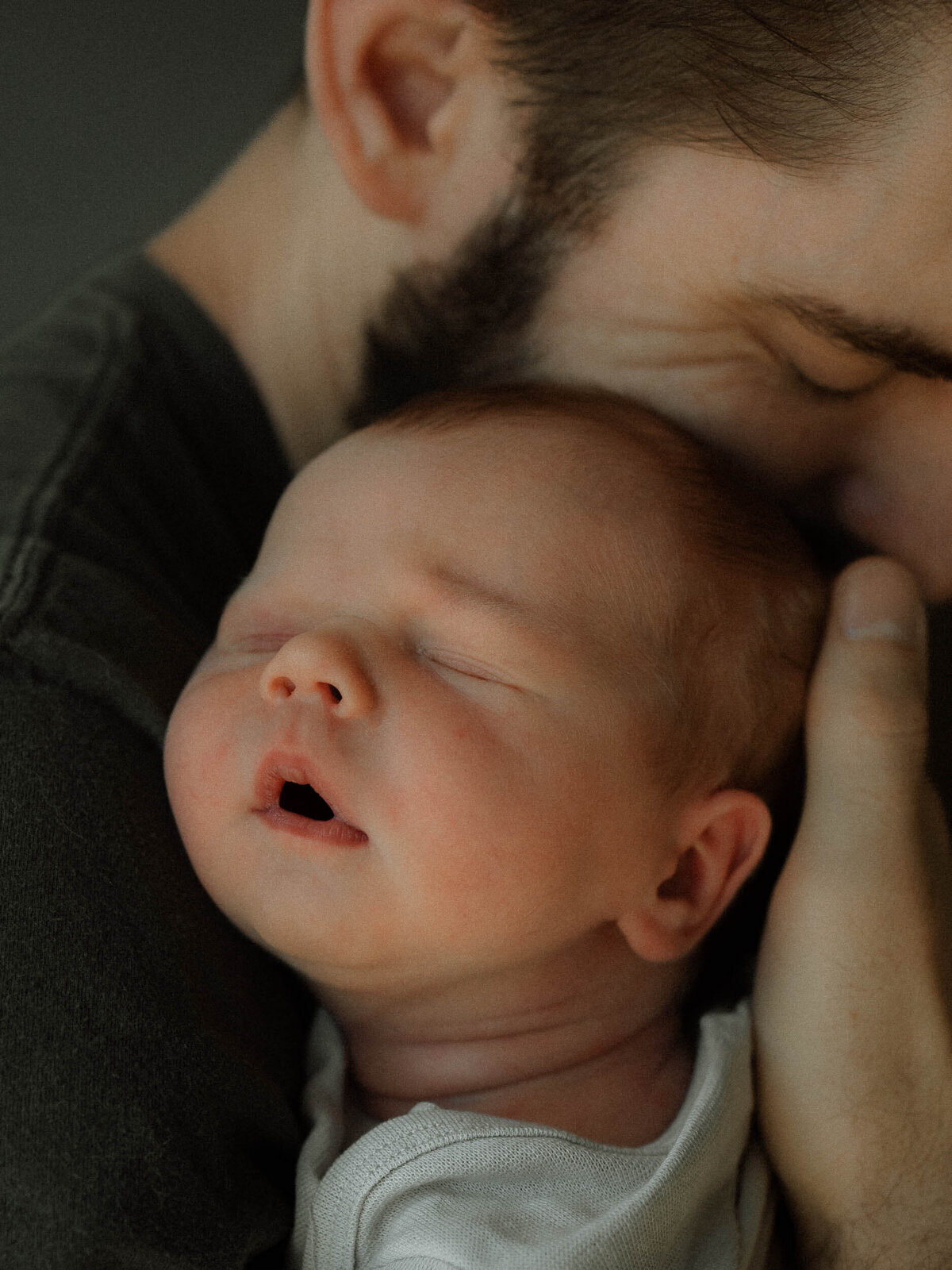 Dad holds his newborn son over his shoulder facing the light while he sleeps with his little mouth open.