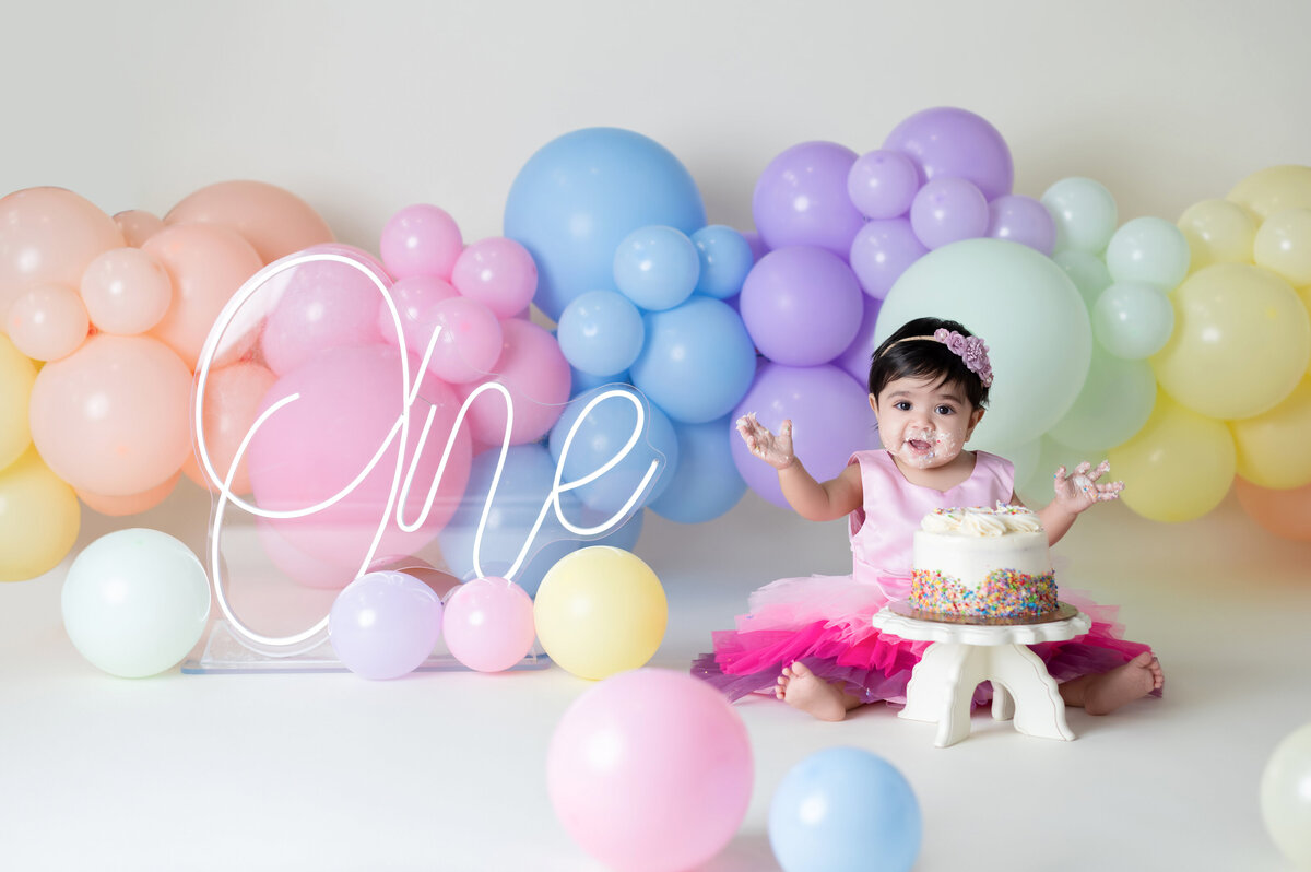 Baby girl in pink tutu makes messy cake, rainbow balloons. Houston cake smash photographer.