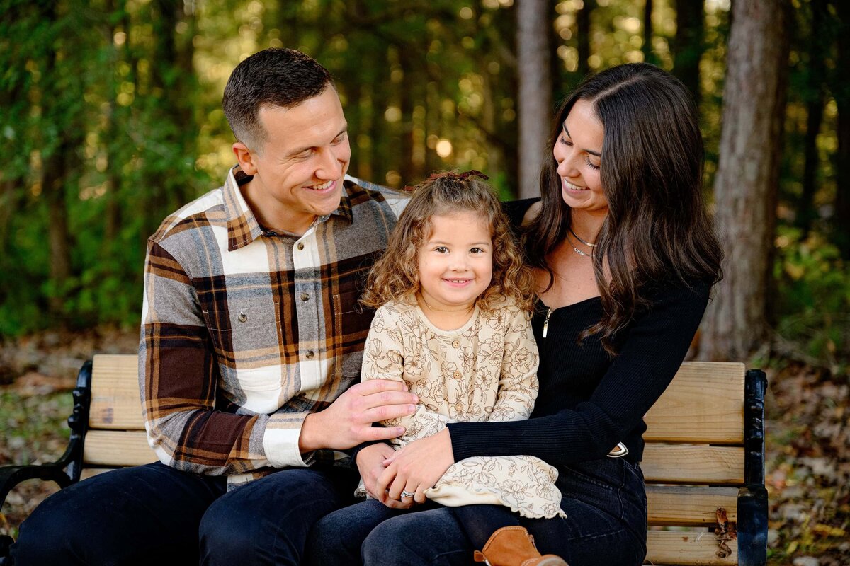 mother and father look down at their little girl while sitting o a bench