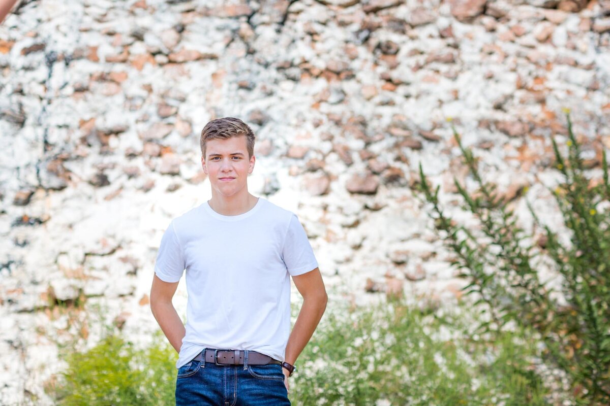 Senior portrait of a boy in a white shirt with green shrubs and a rock wall behind him.